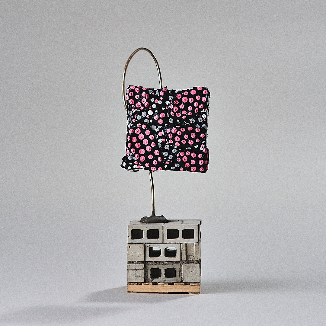 A pink, black and white painted form is held by curved silver wire over miniature cinder blocks.