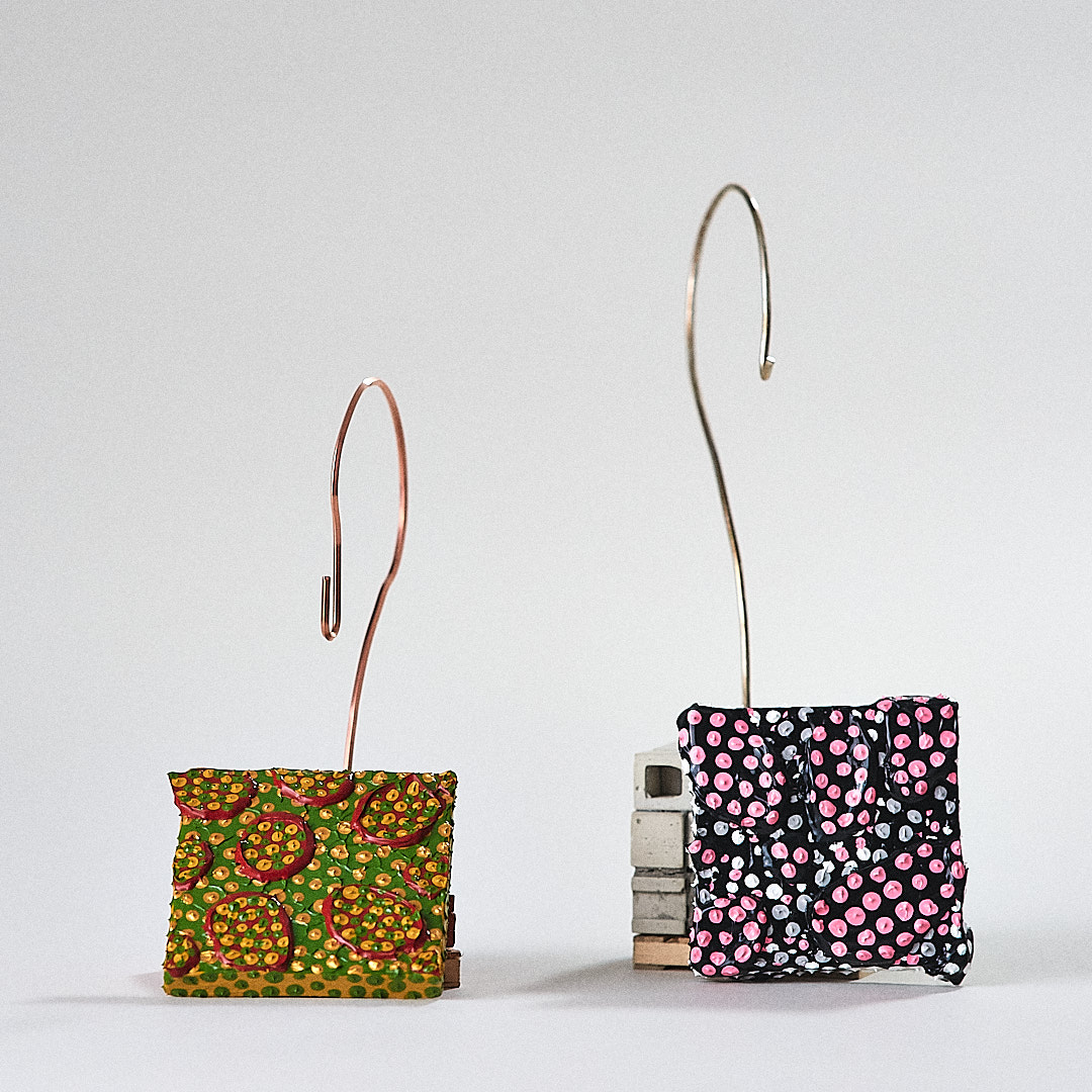 Two patterned sculptural forms suspended by curved wire beside miniature architectural bases.
